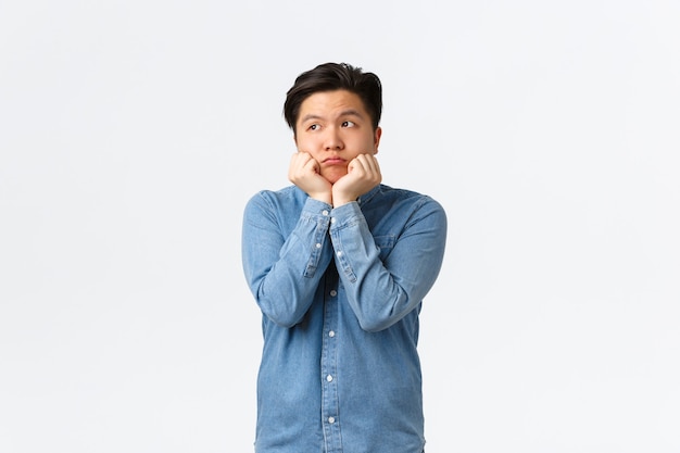 Silly and cute dreamy asian guy looking upper left corner with longing face, leaning on palms timid, thinking about something he desires, feeling bored or sad, standing white background.