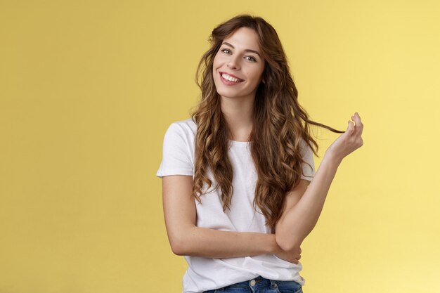 Silly cute feminine sensual curly-haired woman tilt head playing hair strand rolling curl smiling delighted interested listening flirty conversation coquettish gazing camera stand yellow background