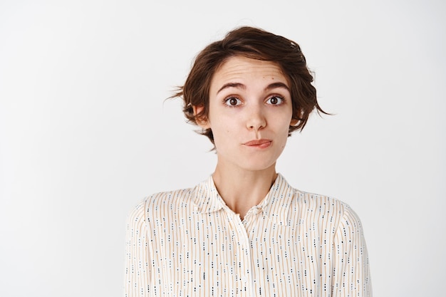 Silly and cute young woman making innocent face, raising eyebrows and smirking, standing on white wall in casual blouse