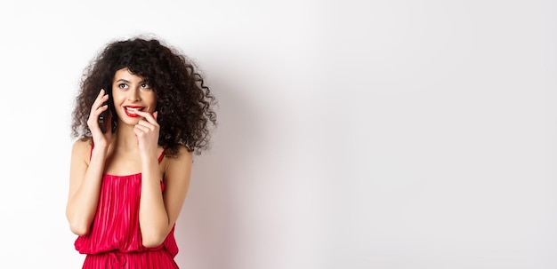 Silly fashionable woman in red dress and lips biting fingernail duing phone call thinking on mobile