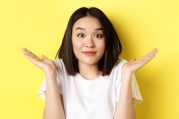 Free photo silly mistake. close up of cute asian girl saying sorry, shrugging shoulders and smiling with oops face expression, standing over yellow background