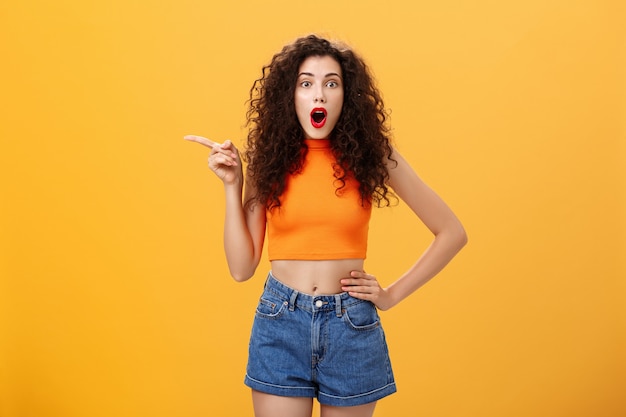 Silly and moody european party girl with curly hairstyle in red lipstic and stylish orange cropped top complaining on bad weather raising hands pointing and looking up with displeased unhappy face