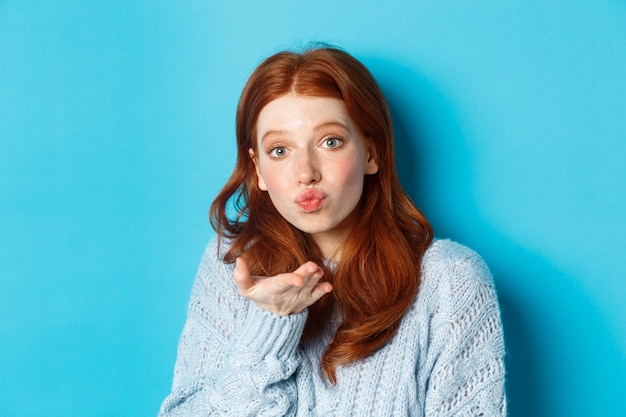 Silly redhead woman in sweater, blowing air kiss at camera with puckered lips, standing against blue background