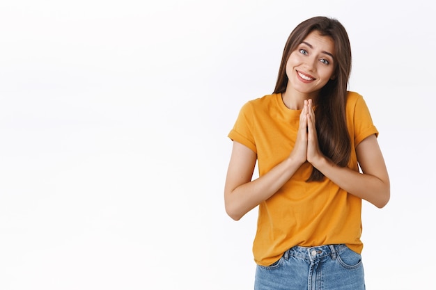 Silly, smiling clingy girl with angel expression, hold hands in pray, tilt head and coquettish gazing camera, begging for favor, lend money from friend, asking advice, apologizing, white background