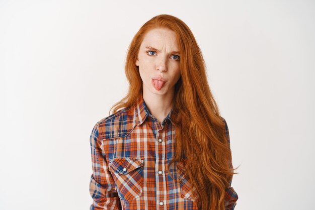 Глупая девочка-подросток с рыжими волосами хмурится и показывает надутый язык, разочарованно стоя на белом фоне