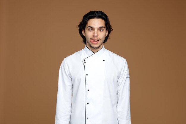 silly young male chef wearing uniform looking at camera with crossed eyes showing tongue isolated on brown background