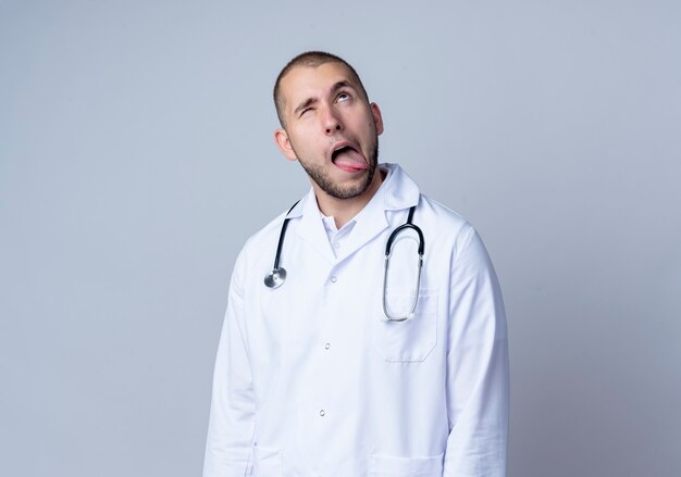 Silly young male doctor wearing medical robe and stethoscope around his neck looking up and showing tongue with one eye closed isolated on white wall