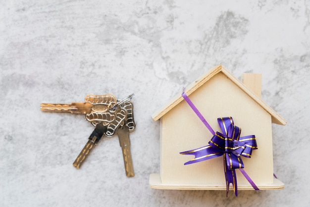 Silver keys near the house wooden model with ribbon bow on white concrete wall