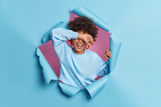 Foto gratuita sincera donna dai capelli ricci copre gli occhi con il braccio risatine nasconde positivamente i sorrisi del viso ampiamente indossa pose casual jumper attraverso il muro di carta blu