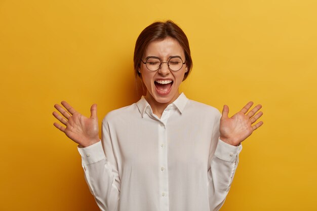 誠実な感情と感情の概念。大喜びの白人女性は、手のひらを上げ、笑い、目を閉じ、面白い冗談を聞き、丸いメガネと白いシャツを着て、黄色い壁にモデルを