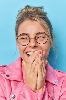 誠実な人間の感情。ポジティブな若い女性が手で口を覆い、積極的に集中して笑い、明るい感じが丸い眼鏡をかけているピンクのジャケットが青い背景に対して屋内でポーズをとる