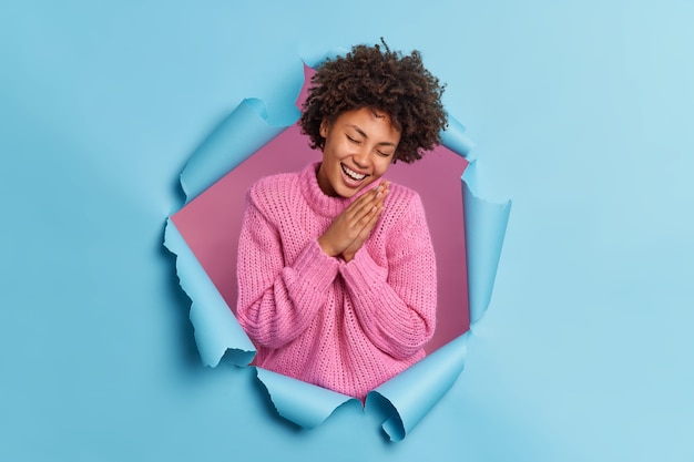 Sincere optimistic curly haired woman keeps palms pressed together chuckles from joy expresses natural emotions wears knitted sweater poses through paper ripped wall