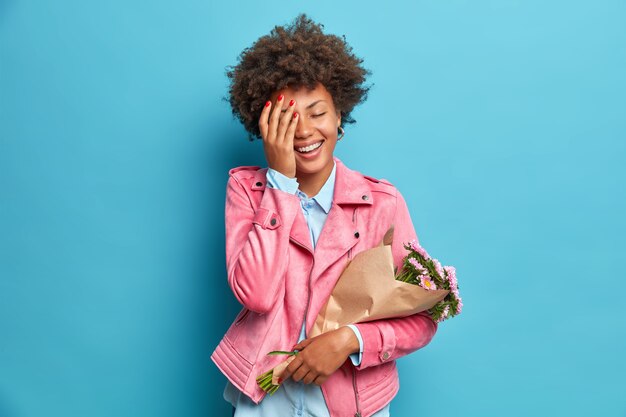 Sincere positive woman dressed in fashionabe clothes feels very happy to get bouquet of flowers from beloved person makes face palm isolated over blue wall