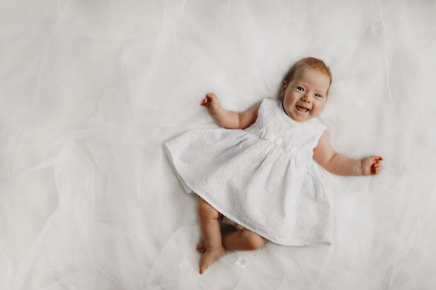 Sincere smile of tiny baby girl dressed in white dress on the bed