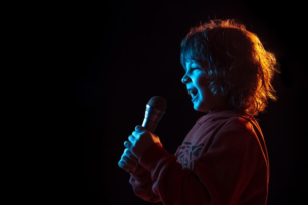 Singing like celebrity, rockstar. Caucasian boy's portrait on dark studio background in neon light. Beautiful curly model. Concept of human emotions, facial expression, sales, ad, music, hobby, dream.