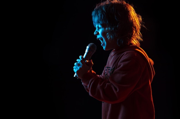 Singing like celebrity, rockstar. Caucasian boy's portrait on dark studio background in neon light. Beautiful curly model.