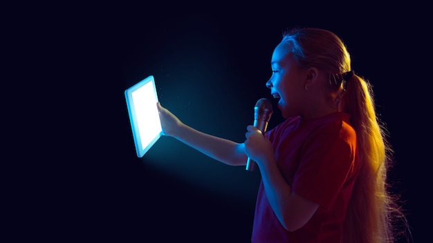 Singing for vlog. Caucasian girl's portrait on dark background in neon light. Beautiful female model using tablet. Concept of human emotions, facial expression, sales, ad, modern tech, gadgets. Flyer.