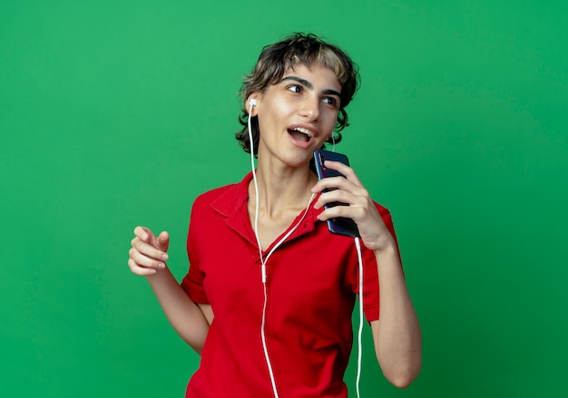 Singing young caucasian girl with pixie haircut wearing earphones and using mobile phone as microphone looking at side