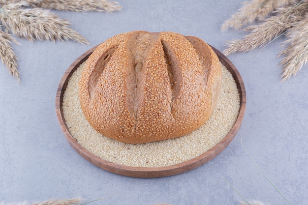 Буханка хлеба на подносе, наполненном семенами кунжута, в окружении сушеных стеблей ковыля на мраморной поверхности
