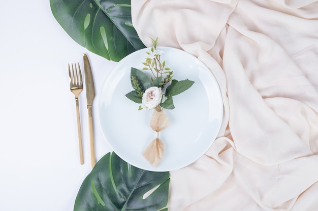 Free photo single rose on white plate with fake leaves and tablecloth.