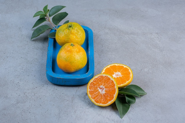 Single tangerine on a platter next to sliced tangerines with leaves on marble background. 