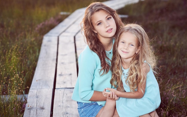 Free photo sisters are relaxing outdoors while sitting on a wooden path in a field at sunset.