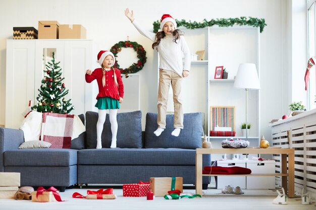 Сестры прыгают и играют на диване, девушки в новогодней шапке