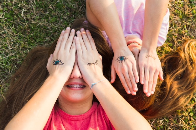 Sisters lying on green grass covering their eyes with tattoos on palm