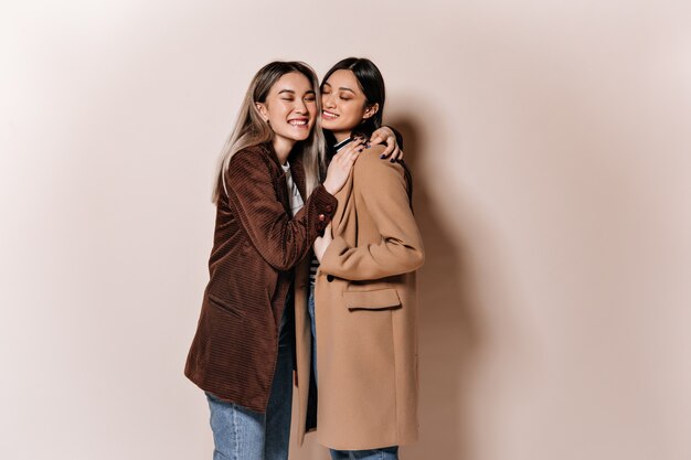 Sisters in stylish outfits posing on beige wall and hugging