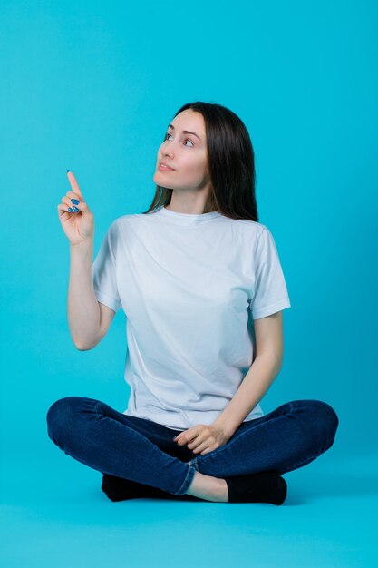 Sitting on floor girl is looking up by pointing left with forefinger on blue background