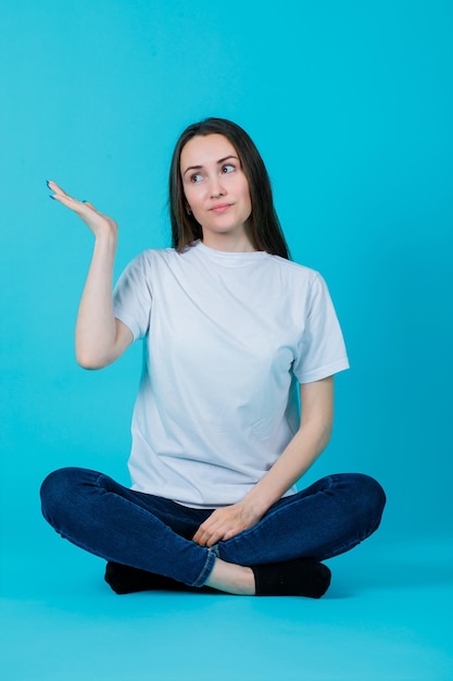 Sitting on floor girl is pointing left with hand on blue background