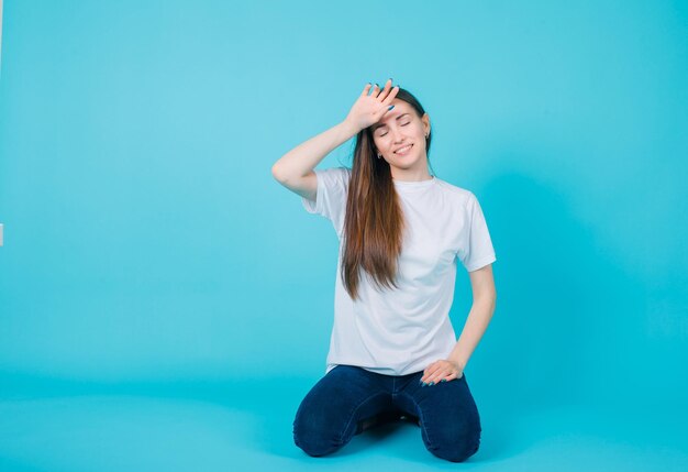 Sitting on floor girl is putting hand on forehead on blue background