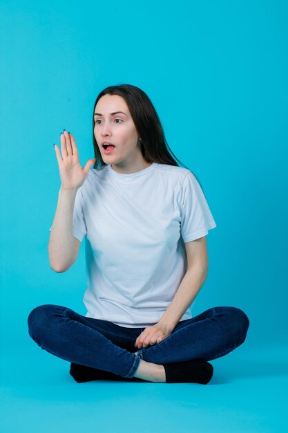 Sitting on floor girl is screaming by holding hand near mouth on blue background