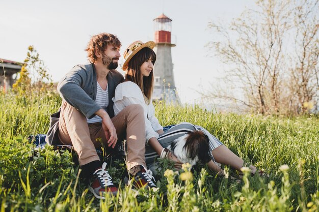 Sitting in grass young stylish hipster couple in love walking with dog in countryside, summer style boho fashion, romantic