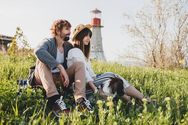 Sitting in grass young stylish hipster couple in love walking with dog in countryside, summer style boho fashion, romantic