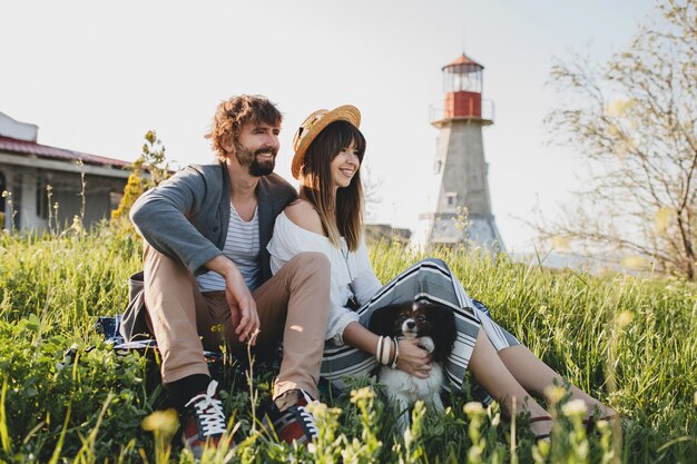 Sitting in grass young stylish hipster couple in love walking with dog in countryside, summer style boho fashion, romantic