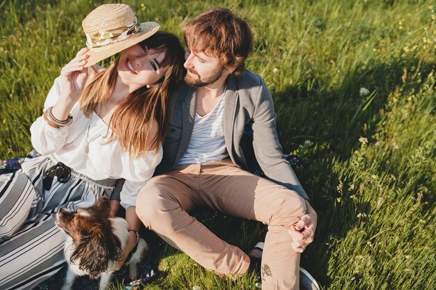 Sitting in grass young stylish hipster couple in love walking with dog in countryside, summer style boho fashion, romantic