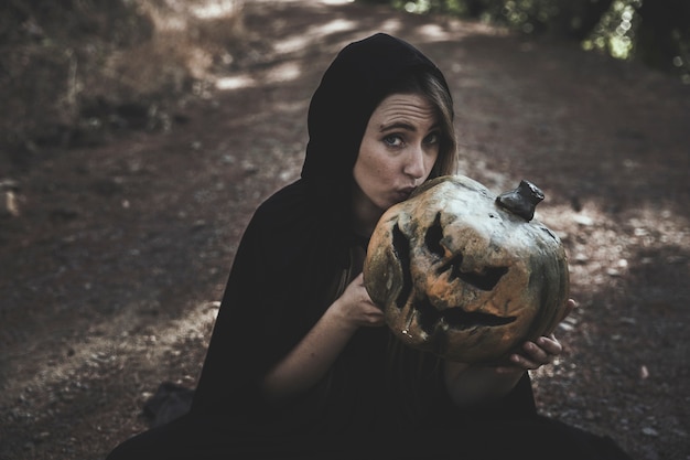 Free photo sitting woman in witch costume kissing frightful pumpkin