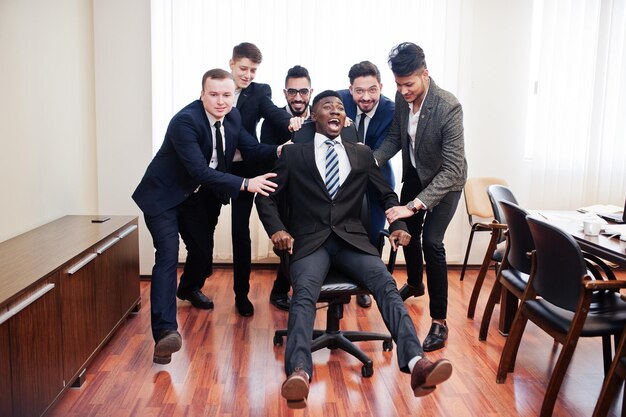 Шесть многорасовых бизнесменов, стоящих в офисе, и катающийся на стуле Разнообразная группа сотрудников-мужчин в формальной одежде, веселящихся