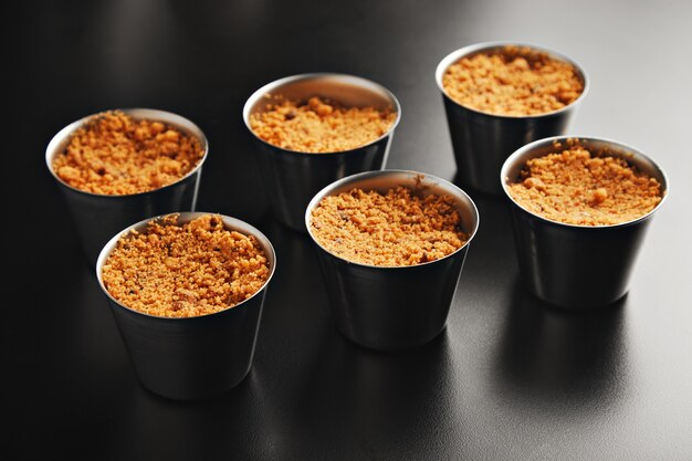 Six portions of apple crumble dessert in individual steel cups on shiny black table