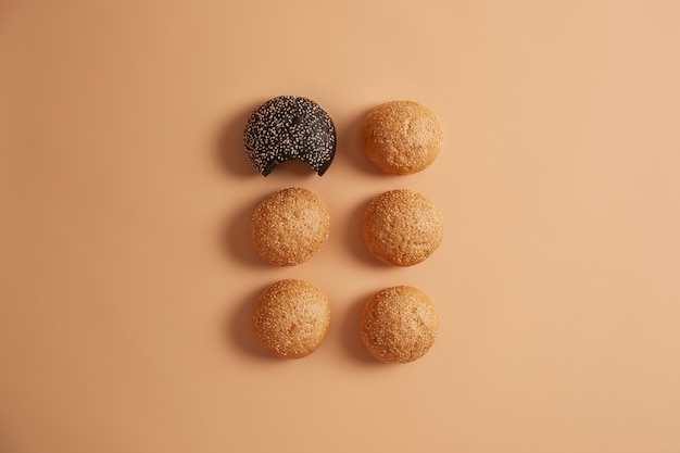 Six small round burger buns made of yeast dough with sesame seeds on beige background. Bakery products for making hamburger. Unhealthy nutrition concept. One black roll is bitten. Fresh food
