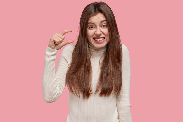 Size matters. Pleased brunette young woman demonstrates very tiny object, smiles positively, wears casual white sweater