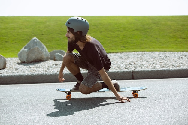 Foto gratuita guidatore di skateboard facendo un trucco sulla strada della città in una giornata di sole. giovane uomo in attrezzatura equitazione e longboard sull'asfalto in azione. concetto di attività per il tempo libero, sport, estremo, hobby e movimento.
