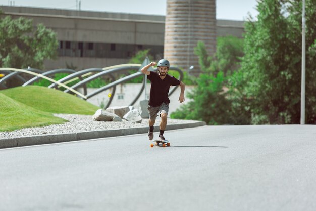 Скейтбордист делает трюк на улице города в солнечный день. Молодой человек в снаряжении верховой езды и лонгбординга на асфальте в действии. Концепция досуга, спорта, экстрима, хобби и движения.