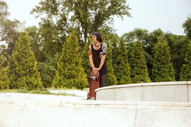 Skateboarder resting after riding at the city's street in cloudly day. Young man in sneakers and cap with a longboard on the asphalt. Concept of leisure activity, sport, extreme, hobby and motion.