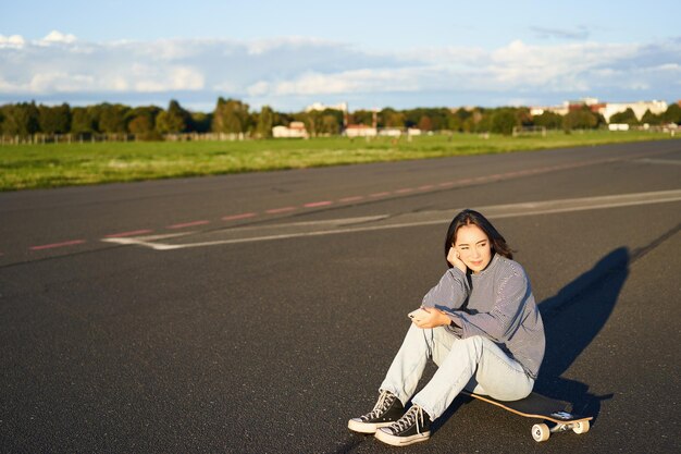 Фигуристка сидит на своем скейтборде на дороге, разговаривая со смартфоном в мобильном приложении