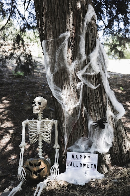 Скелет, держащий тыкву возле таблички Хэллоуина, опираясь на дерево