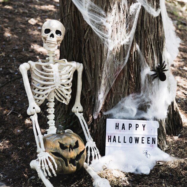 Skeleton sitting near tree with pumpkin and Happy Halloween inscription