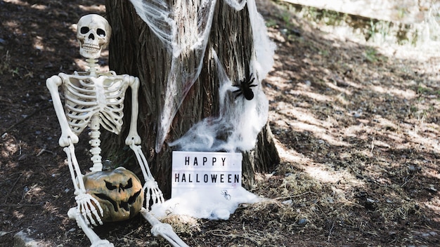 木に傾いているハロウィーンのボードの近くに座ってカボチャとスケルトン