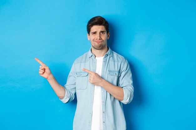 Скептически настроенный взрослый мужчина, указывая пальцами влево, выглядит сомнительно и неуверенно, стоит на синем фоне